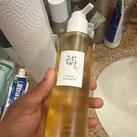 Beauty of Joseon Ginseng Cleansing Oil tenu en main près d’un lavabo et de produits de salle de bain.